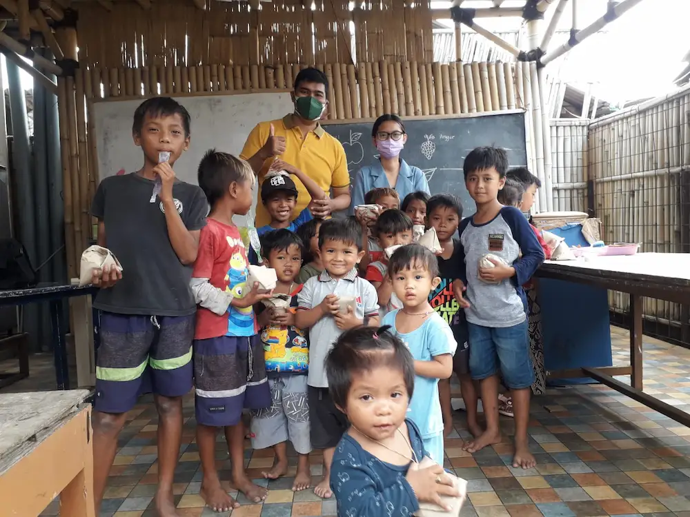 Children gather around two volunteer teachers in a bamboo classroom at our community learning center.