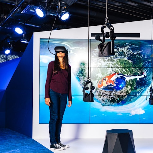 Person wearing a VR headset in front of a large screen showing an aerial view of a helicopter and landscape, surrounded by VR equipment.