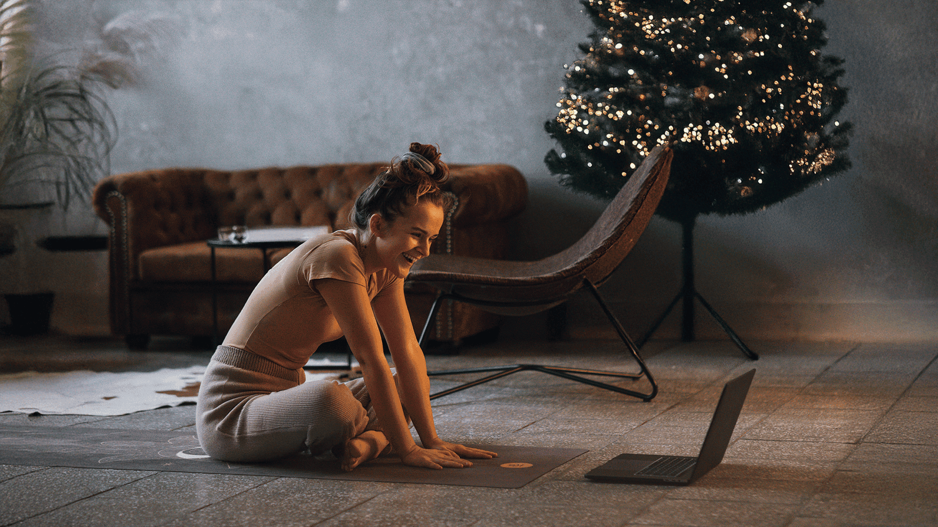 Frau sitzt lächelnd auf einer Yogamatte vor einem Laptop, Weihnachtsbaum im Hintergrund.