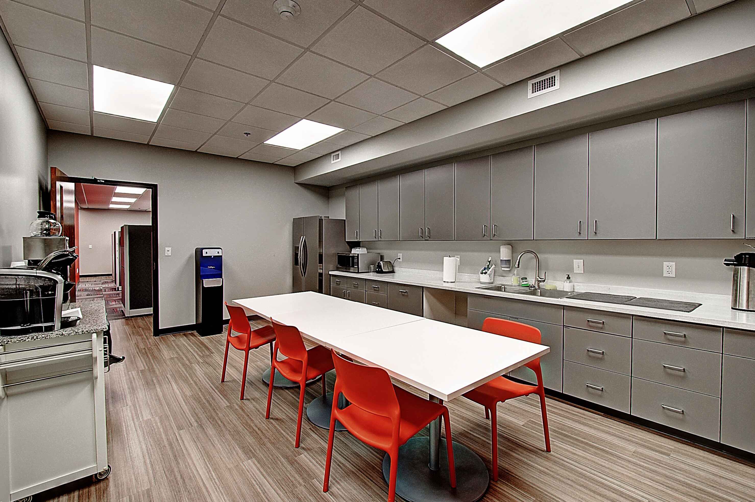 Modern kitchen with gray cabinets, a white table, and red chairs under bright ceiling lights.