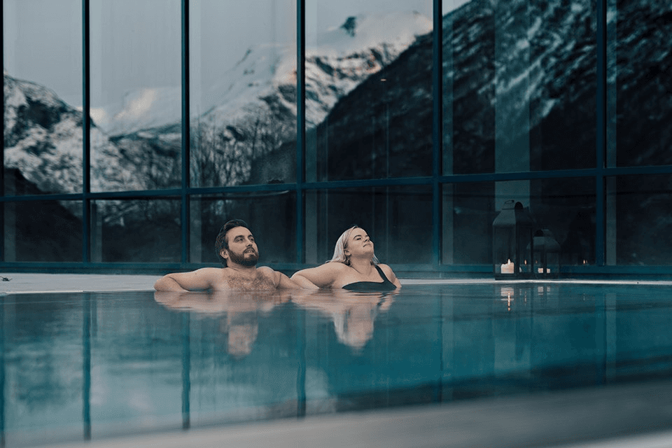 A couple relaxes in a luxurious indoor pool surrounded by large glass windows showcasing a breathtaking snow-covered mountain view, creating a serene and intimate ambiance.