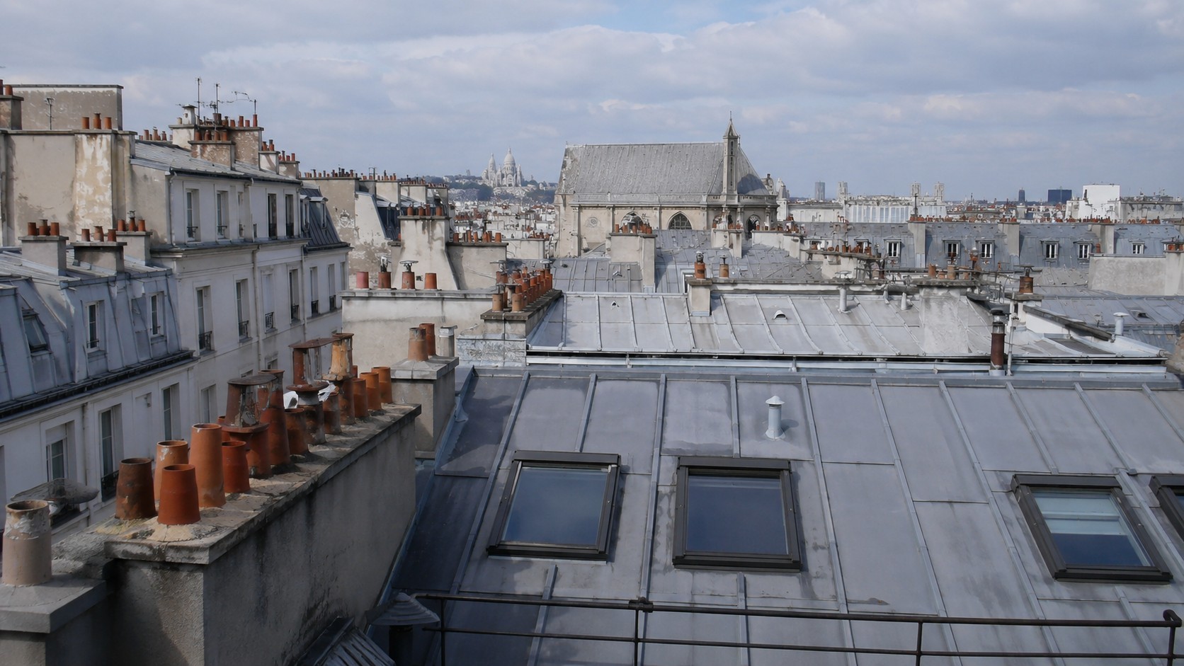 Toiture parisienne en zinc avec rangées de cheminées traditionnelles, fenêtres de toit et immeubles haussmanniens. Vue authentique des toits de Paris prise en hauteur, montrant l’architecture caractéristique de la capitale française et le patrimoine des couvertures en zinc.