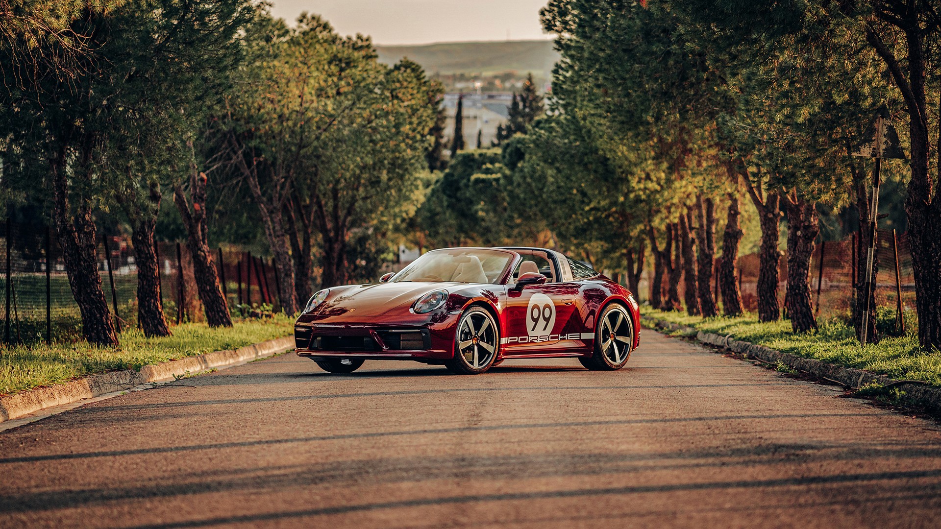 Red Porsche 911 Targa 4S Heritage Design Edition parked on a tree-lined avenue at golden hour, roof down, wearing white "99" race roundels and gold "PORSCHE" side script, with cypress trees and hills behind.