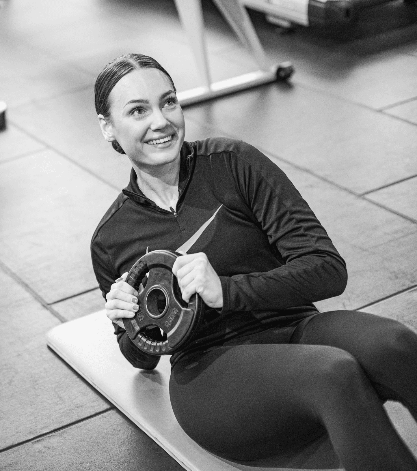 A woman is sitting on a yoga mat and smiling while holding a weight plate during an exercise.