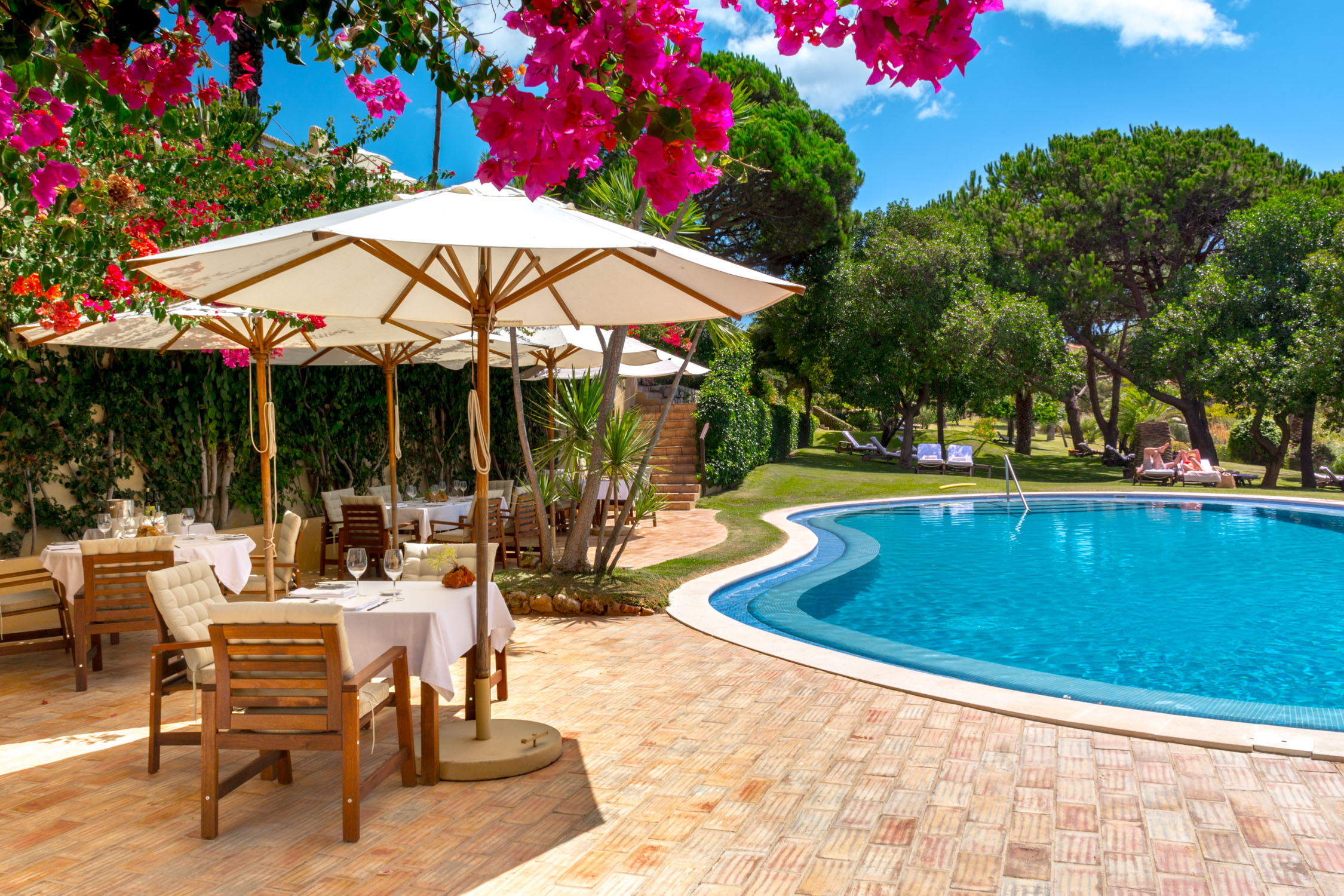 A poolside dining area with wooden tables and chairs under white umbrellas framed by pink bougainvillea flowers.