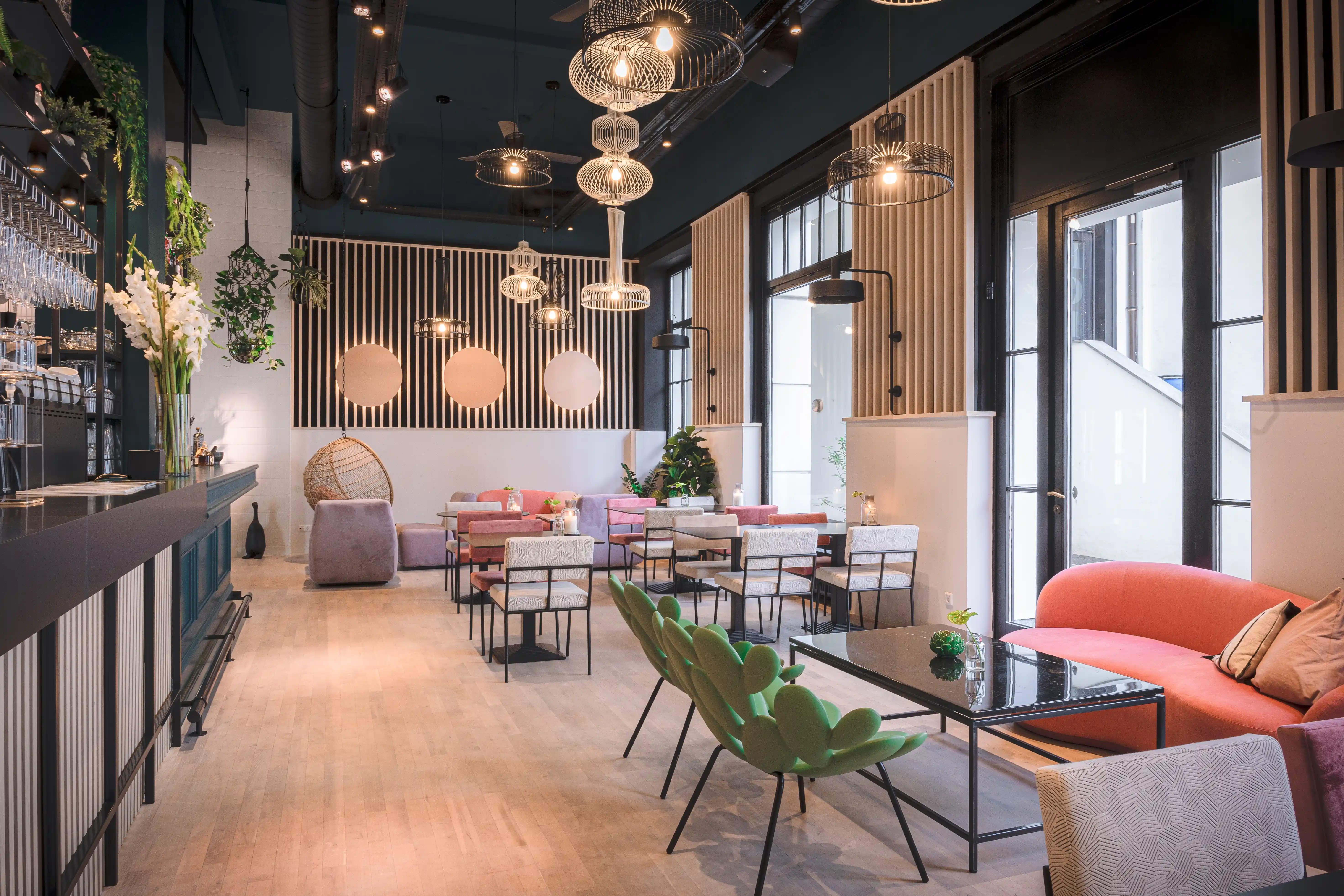 Interior of LOKAL VIER with pink and lilac sofas, green sculptural lounge chairs, woven wire pendant lamps, peach wall circles and a long dark bar counter.