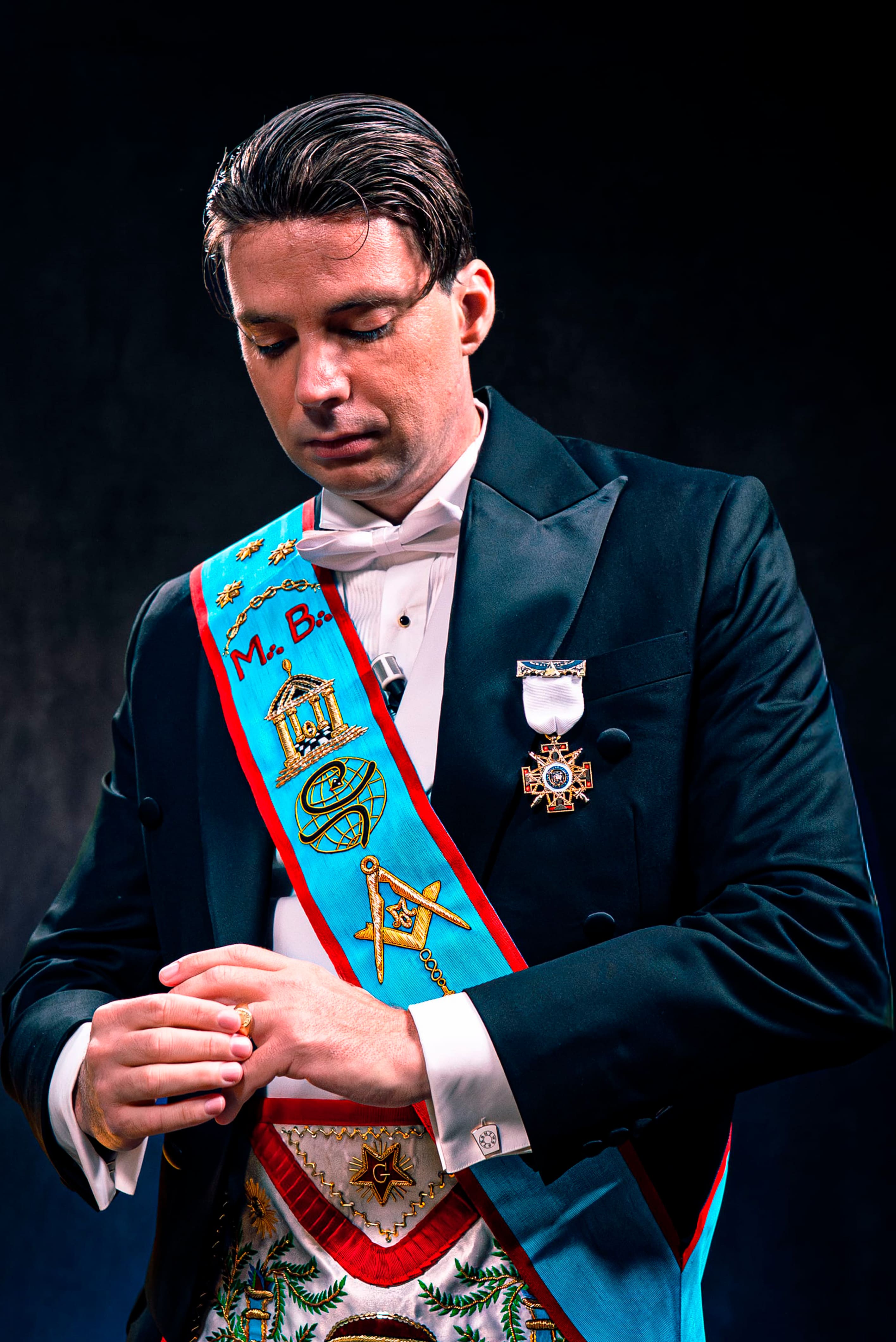 Photographie de portrait professionnelle d'un homme en tenue de Franc-Maçonnerie, portant un smoking noir, un nœud papillon blanc et des insignes. L'homme est vu de face, les yeux baissés et les mains ajustant une bague. L'accent est mis sur l'écharpe d'officier bleu vif (sautoir) ornée de symboles maçonniques (équerre et compas, temple). Le fond sombre et l'éclairage dramatique créent une atmosphère sérieuse et mystérieuse. Cette photo de haute qualité, optimisée par Nux-Studio, est un exemple de production photo pour des interviews sur l'histoire, les organisations secrètes ou des sujets de société (Franc-Maçonnerie).