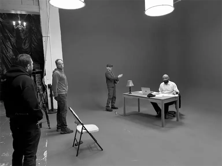 Two focused men in work aprons lean over large blueprints in a dimly lit workshop. A beam of light highlights the papers, creating a dramatic atmosphere.