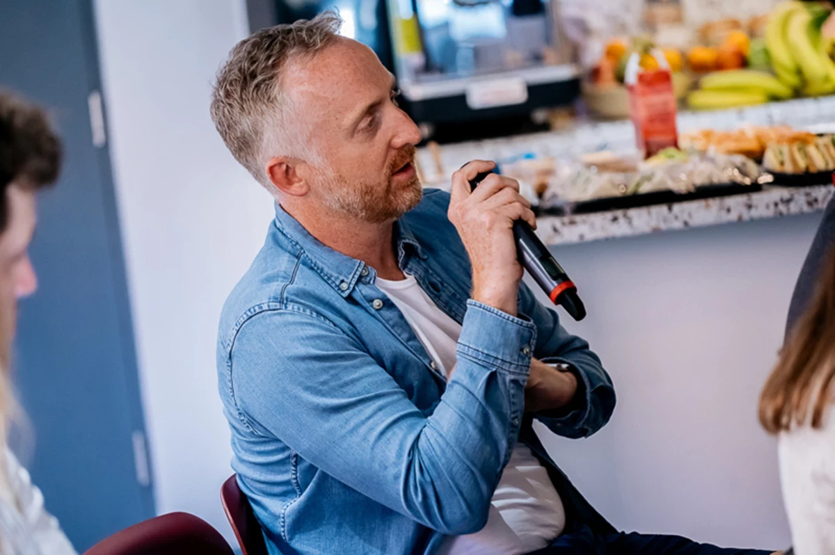 Person in a light denim shirt holding a microphone while seated, with a countertop in the background displaying fruit and snacks.