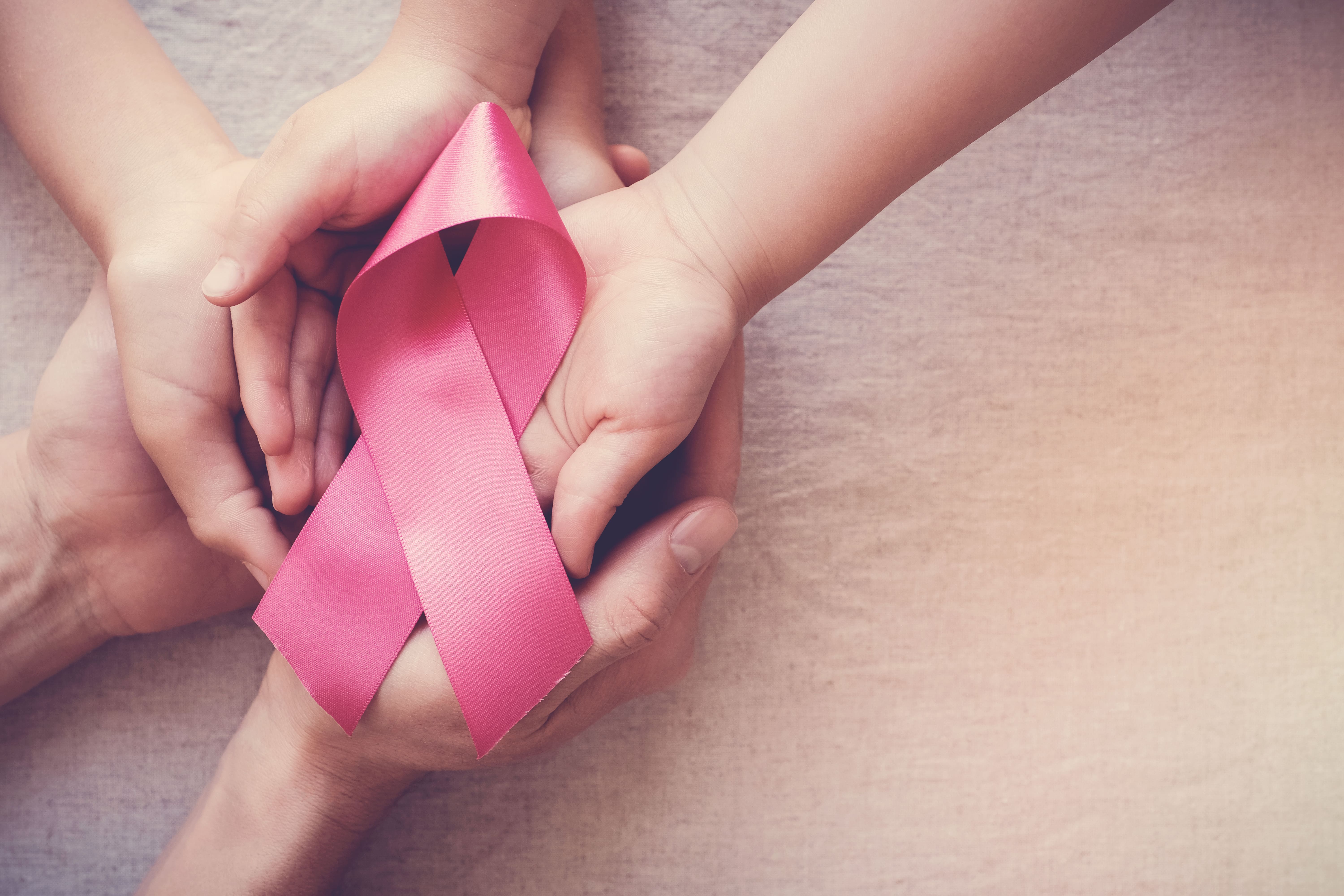 Imagem de duas mãos adultas segurando as pequenas mãos de uma criança, ambas apoiando um laço rosa, símbolo internacional de conscientização e apoio à luta contra o câncer de mama. O fundo é neutro e suave, realçando a mensagem de cuidado, proteção e solidariedade que o gesto transmite.