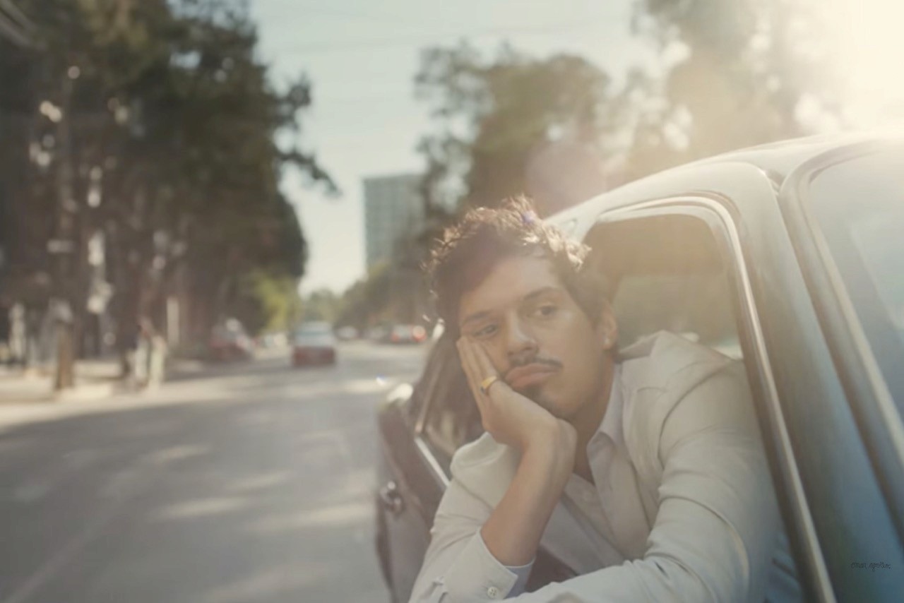 Omar Apollo leans out of a car window on a sunny street, gazing thoughtfully into the distance. The scene conveys a sense of relaxation and daydreaming.