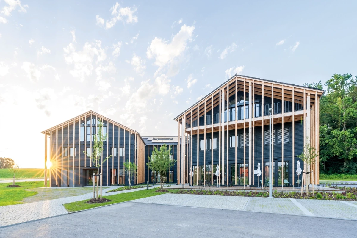 Moderne Holzkonstruktion mit dunkler Verkleidung und großen Fenstern, umgeben von Grün, gegen einen hellen Himmel.