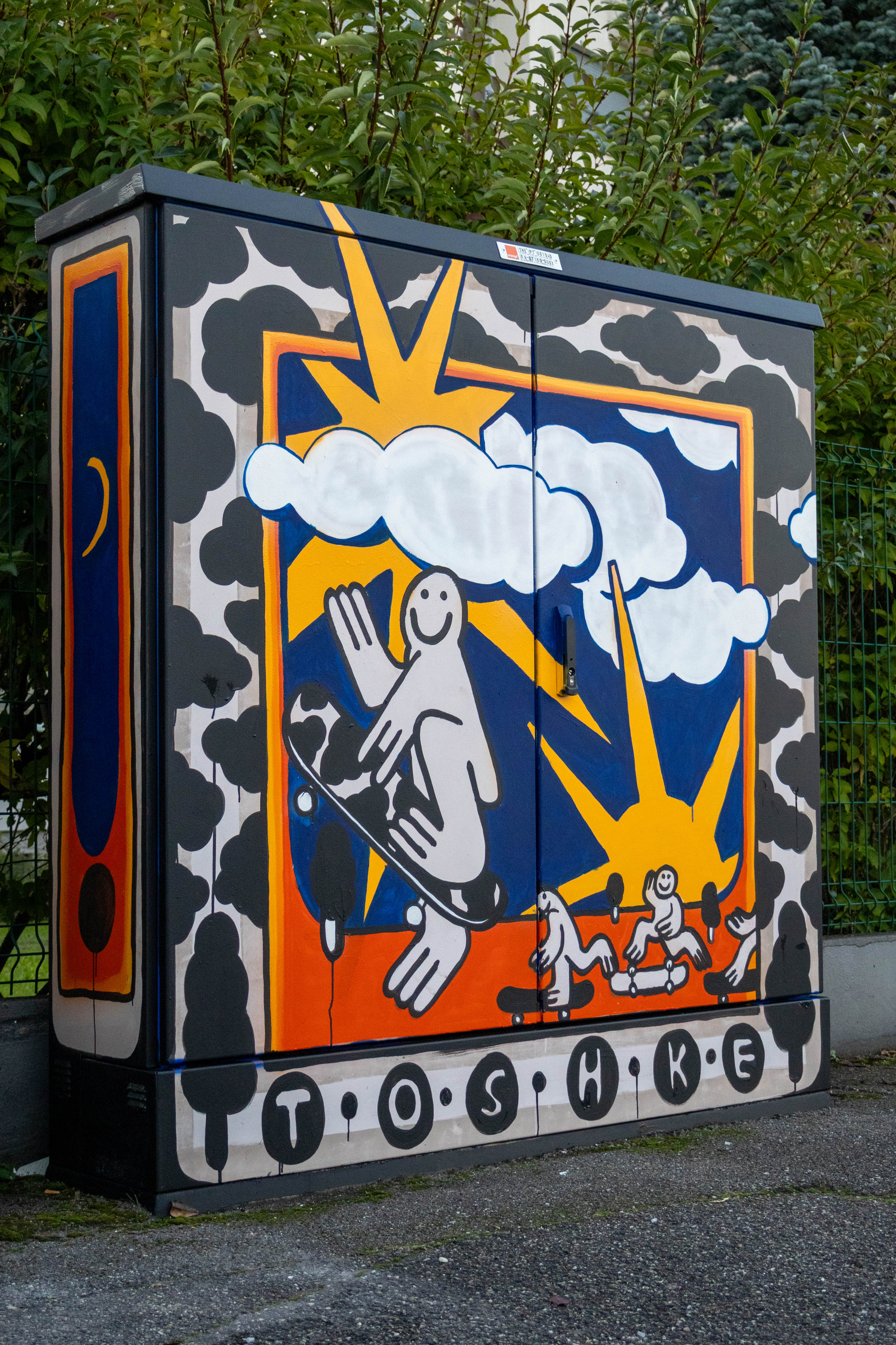 A painted electrical box with joyful black-and-white cartoon characters skating on an orange floor under yellow suns and clouds, with “TOSHKE” at the bottom and an abstract moon pattern on the side.