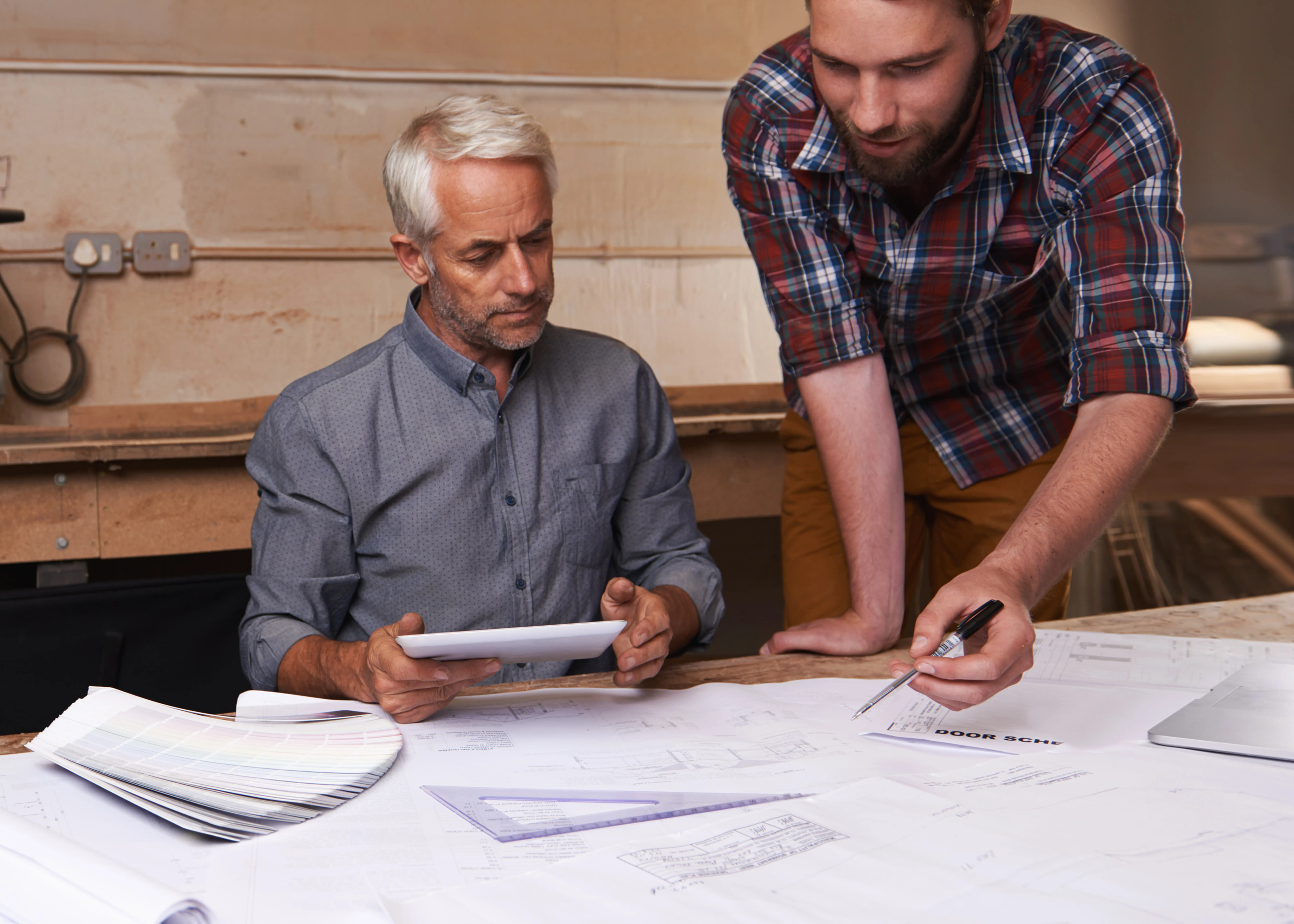 Two professional male designers collaborating over large architectural blueprints and a digital tablet in a studio.
