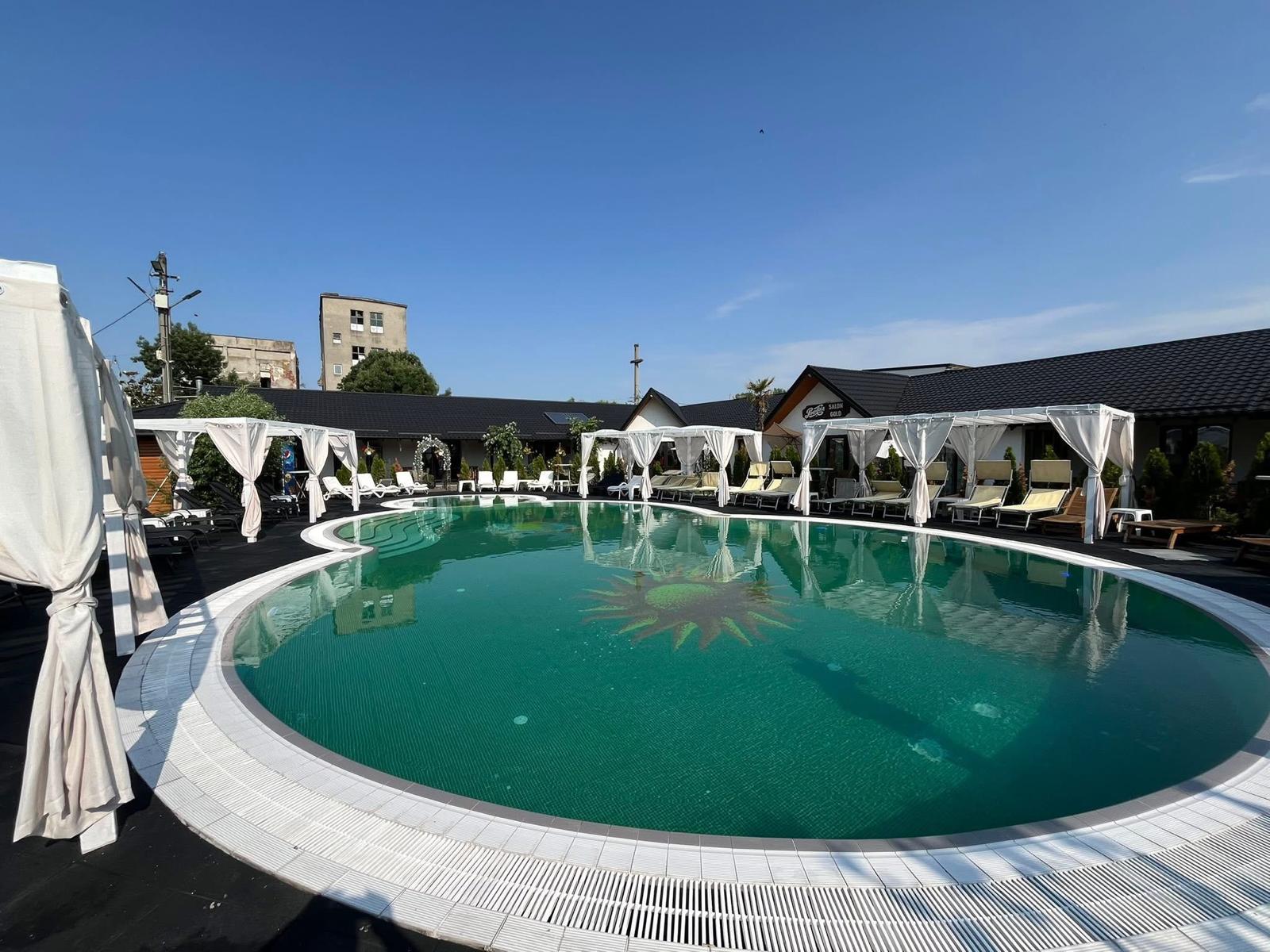A modern, glass-walled house with warm interior lighting is perched on a rocky cliff overlooking a calm sea at sunset, under a clear blue sky.