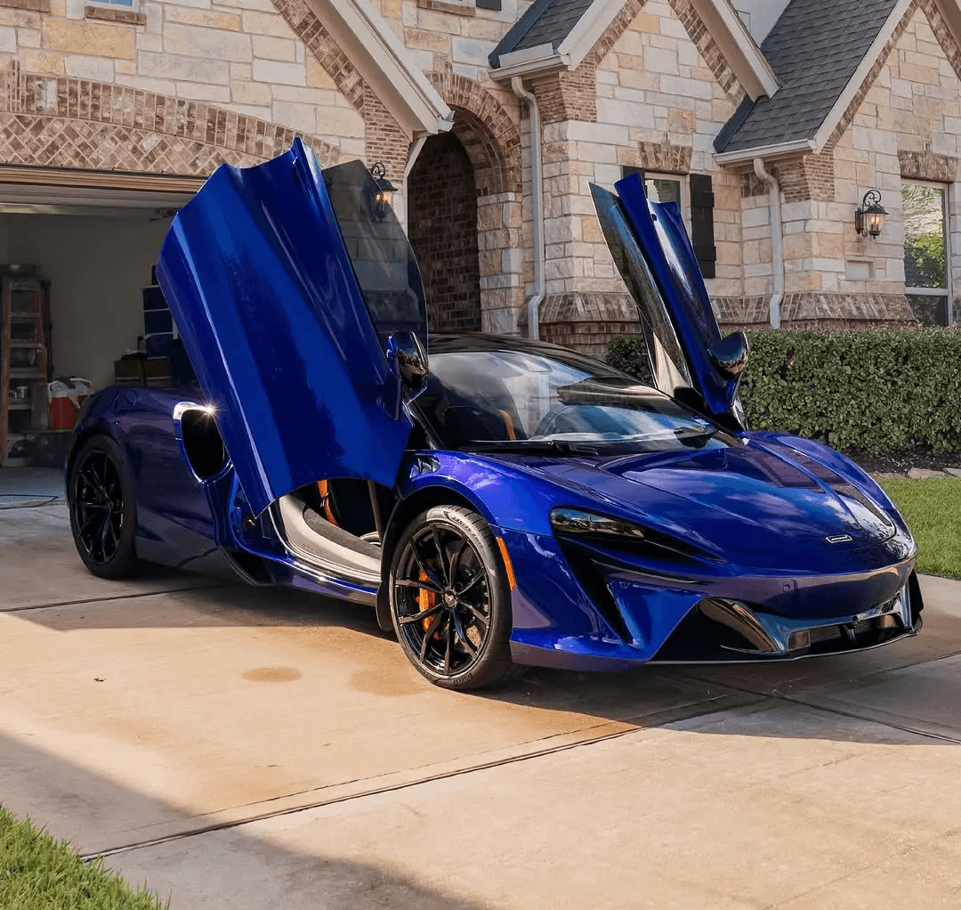 A metallic blue McLaren Artura with its dihedral doors open, parked on a residential driveway in front of a large, luxury stone house.