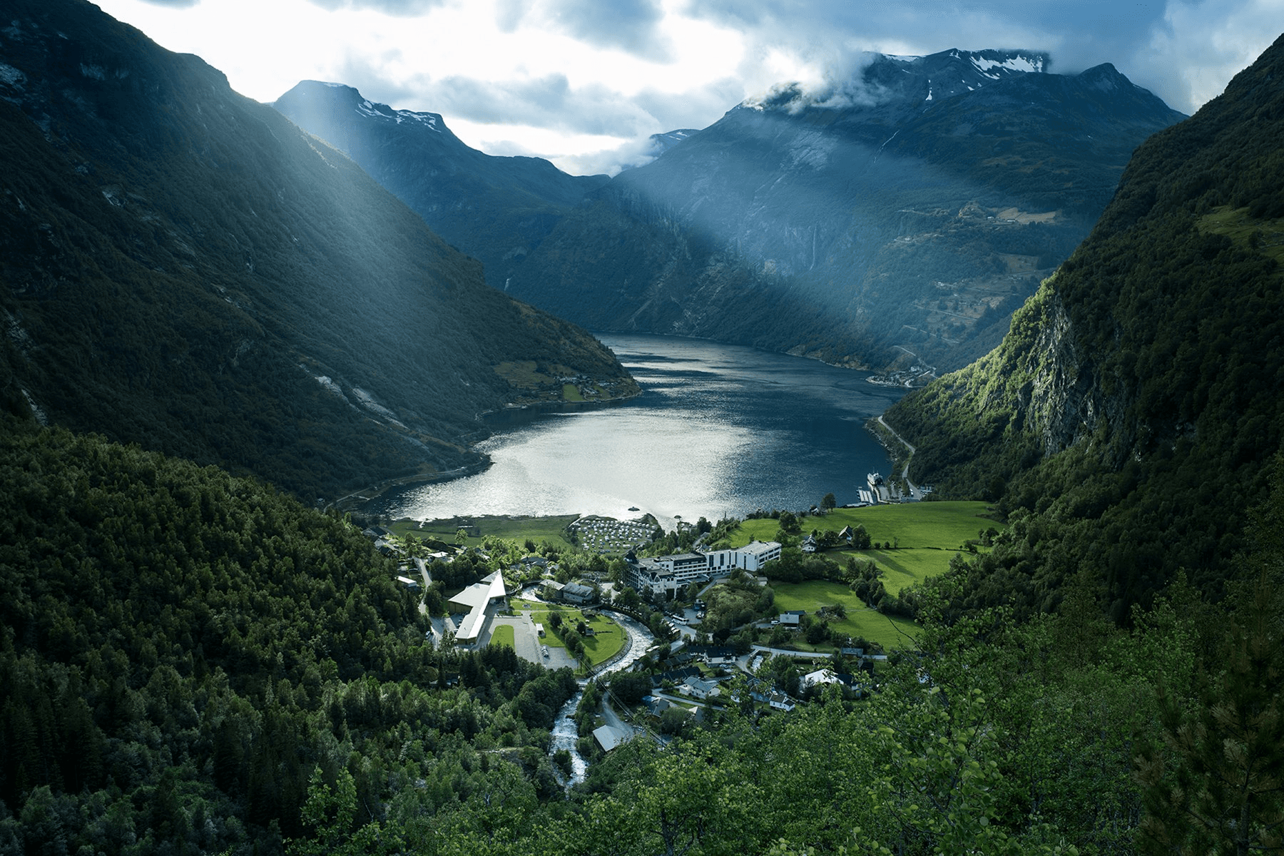 A breathtaking view of a serene valley with a shimmering lake surrounded by majestic mountains, lush greenery, and scattered buildings under a cloudy sky with rays of sunlight breaking through.