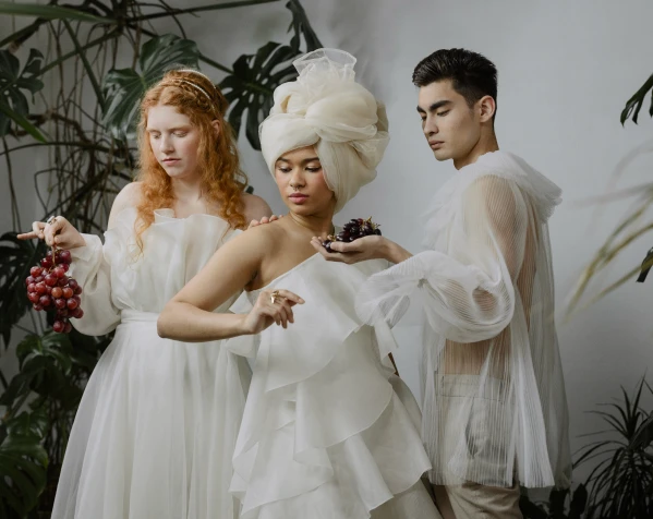 Three people in flowing white garments and elaborate headpieces are posing with bunches of grapes, surrounded by tropical plants.