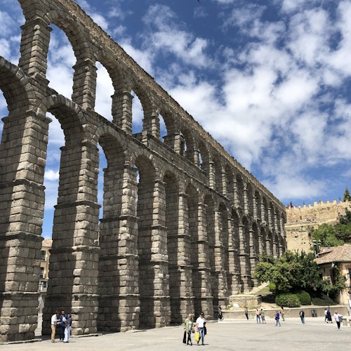 Ancient stone aqueduct with multiple arches under a partly cloudy sky, surrounded by people and buildings.