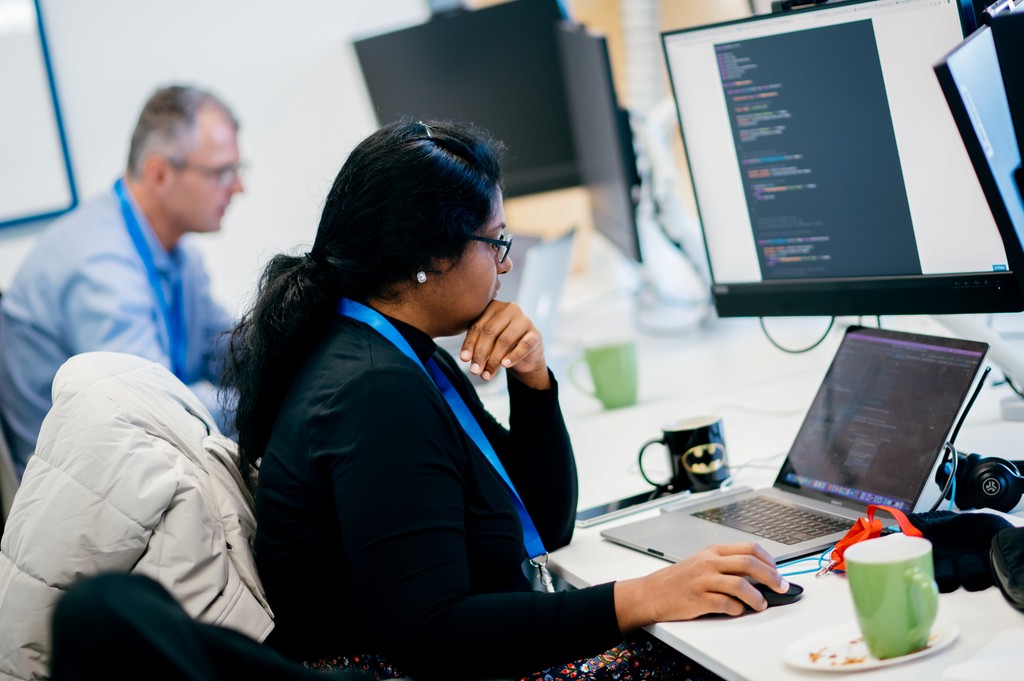 Woman with glasses working on code at a computer, laptop open, another person in background.