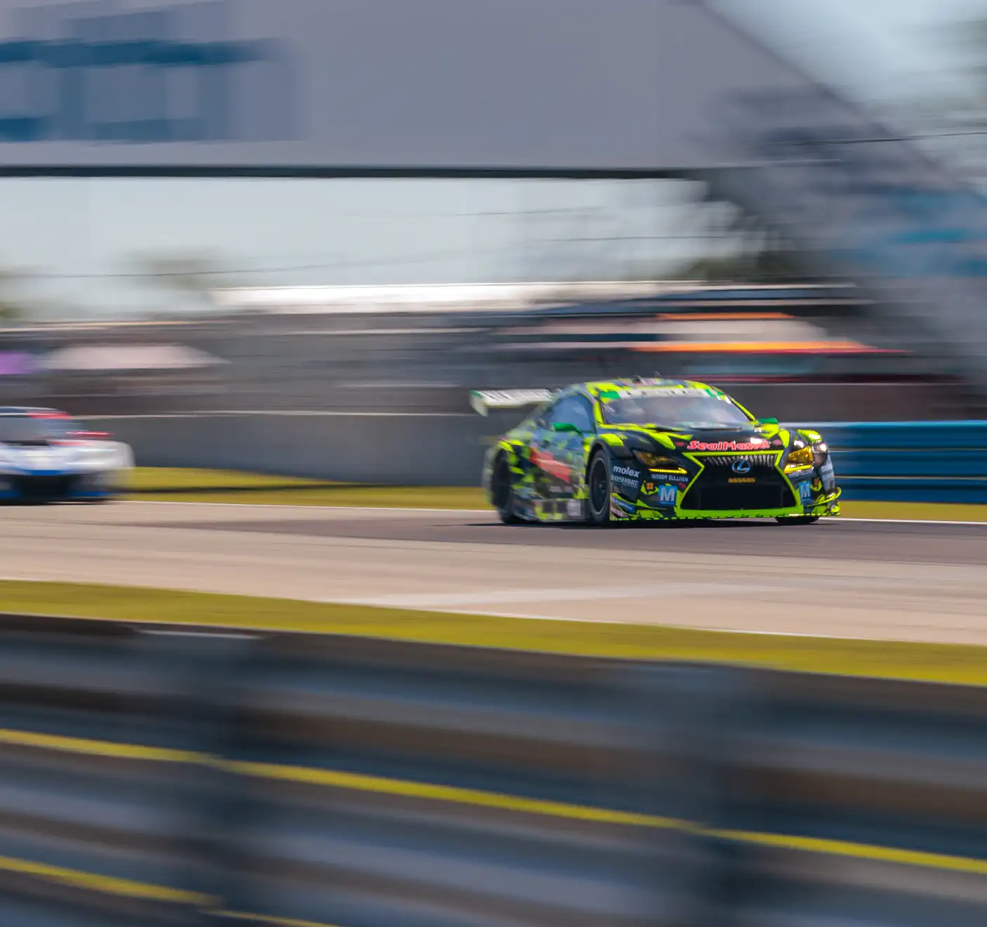 A green and yellow race car speeds on a track, closely followed by a white car, with a blurred banner and trees in the background.