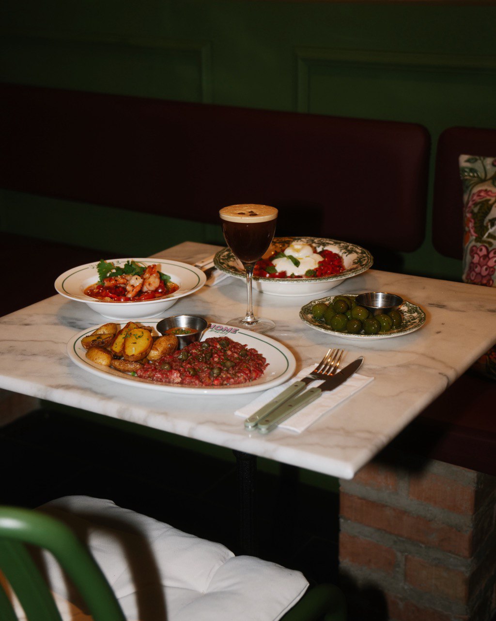 A small round white marble table set with plates of savory dishes, including grilled prawns, steak tartare, roasted potatoes, and green olives, alongside a frothy iced coffee in an elegant glass, situated in a cozy restaurant setting.