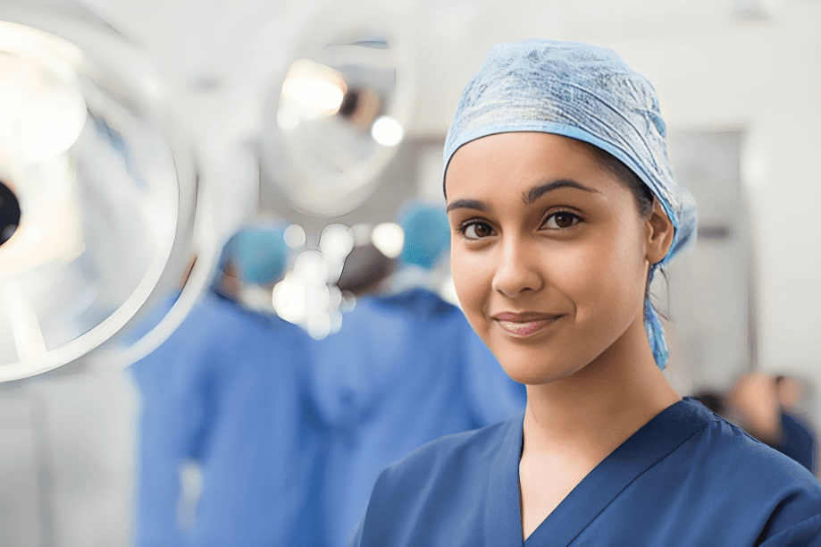 A young female healthcare professional in blue scrubs and a surgical cap smiles confidently, standing in a bright hospital environment with colleagues working in the background.