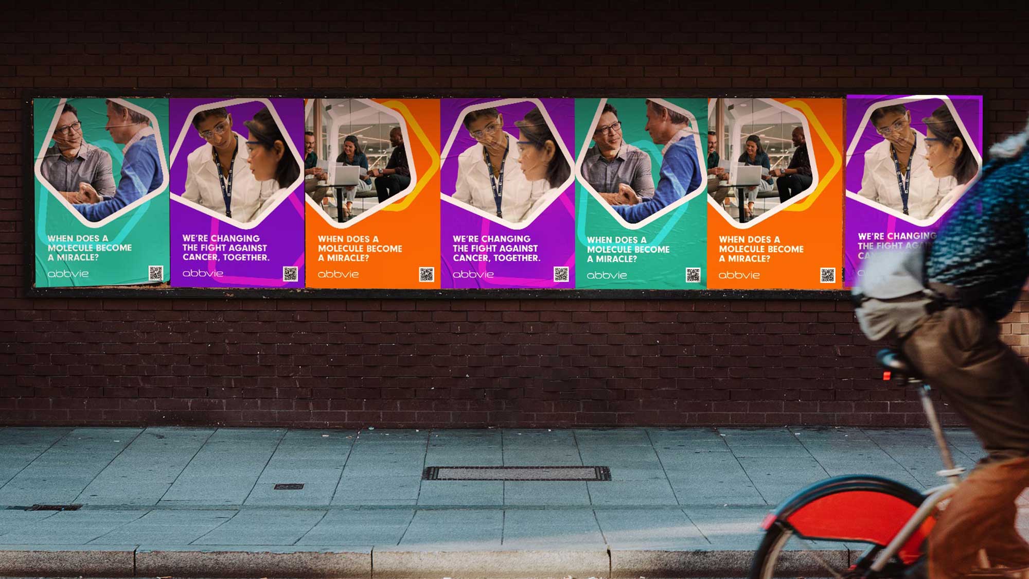 A row of colourful AbbVie recruitment posters pasted along a brick wall as a cyclist rides past