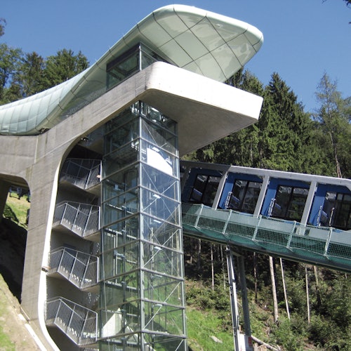 Uma ferrovia funicular moderna com uma estrutura de escadaria de vidro e um trem inclinado passando por uma encosta arborizada.