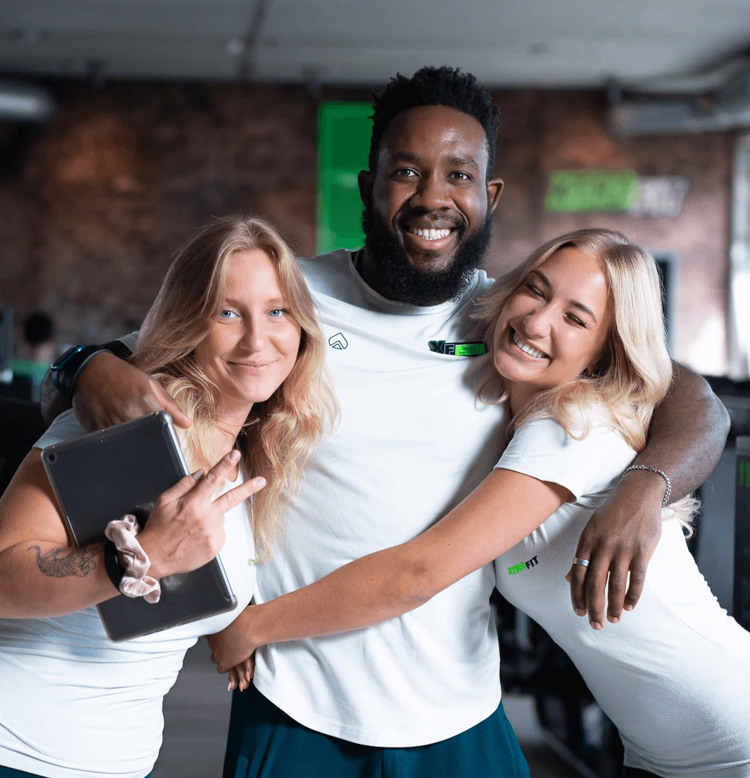 Drei XTRAFIT-Trainer stehen locker beisammen im Cardio-Bereich eines XTRAFIT-Studios. Der Trainer in der Mitte schaut auf einen Tabletcomputer.