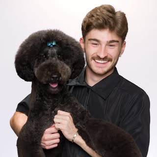 Portrait of Logan Rago holding a stylish black poodle with a blue bow to convey trust, care, and refined grooming expertise.