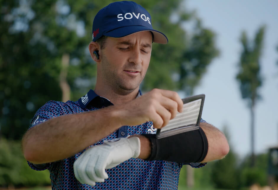 A golfer wearing a glove and cap looks at a scorecard or yardage book attached to his arm on a golf course.