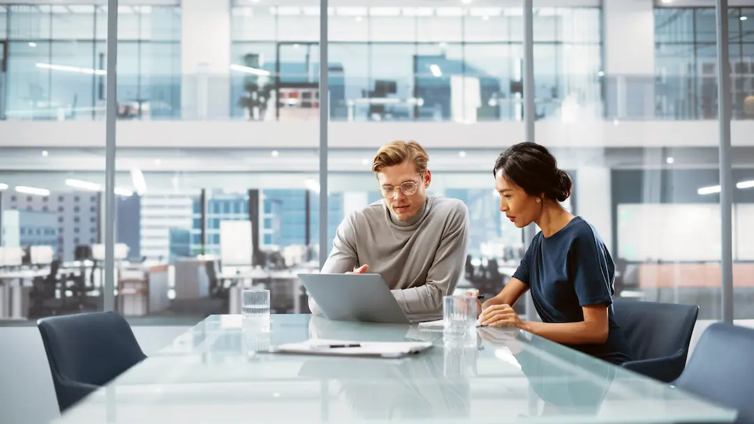 Two colleagues collaborating over a laptop in a modern glass-walled office