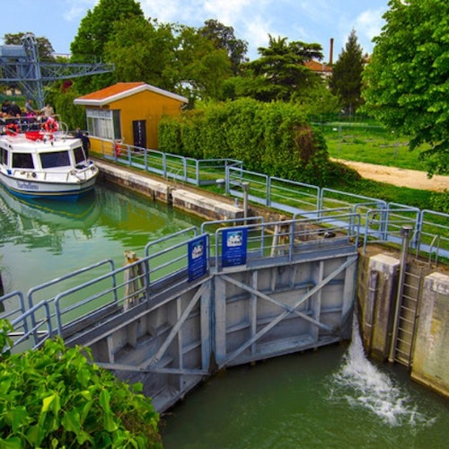 Een boot met passagiers vaart een sluis binnen, omringd door groene bomen en een geel gebouw.