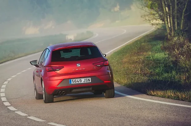 Une SEAT Leon rouge photographiée de dos sur une route sinueuse entourée de verdure. La voiture s’éloigne dans un décor matinal légèrement brumeux, mettant en avant son dynamisme et son allure élégante en pleine conduite.