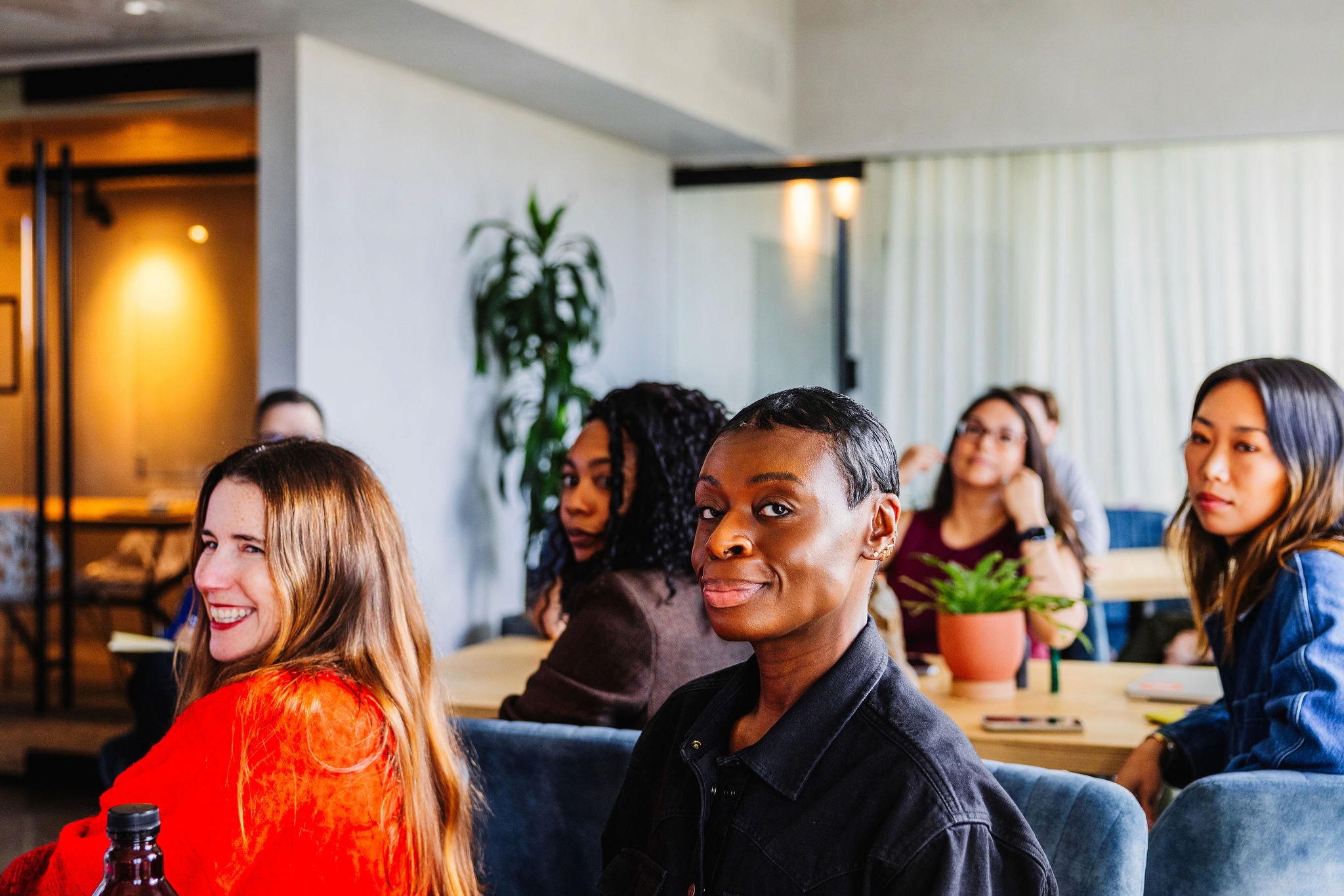 People seated at tables looking toward the camera during a meeting, with notebooks and plants visible in a modern office.
