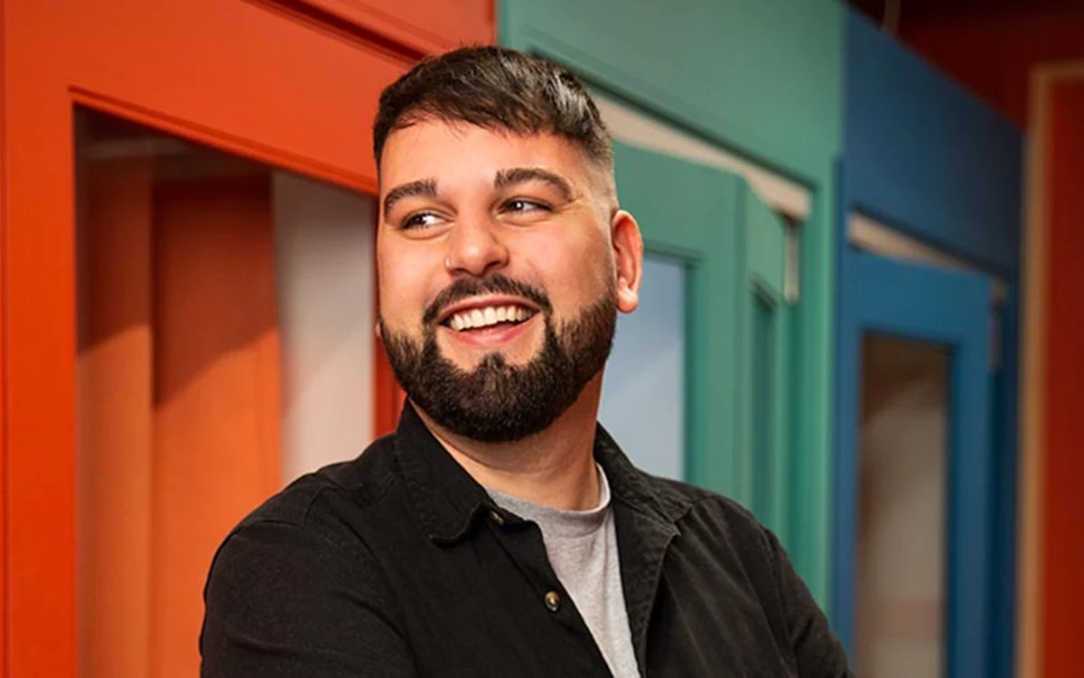 Person wearing a black button-up shirt over a light gray t-shirt, standing in front of brightly colored door frames in orange, teal, and blue.
