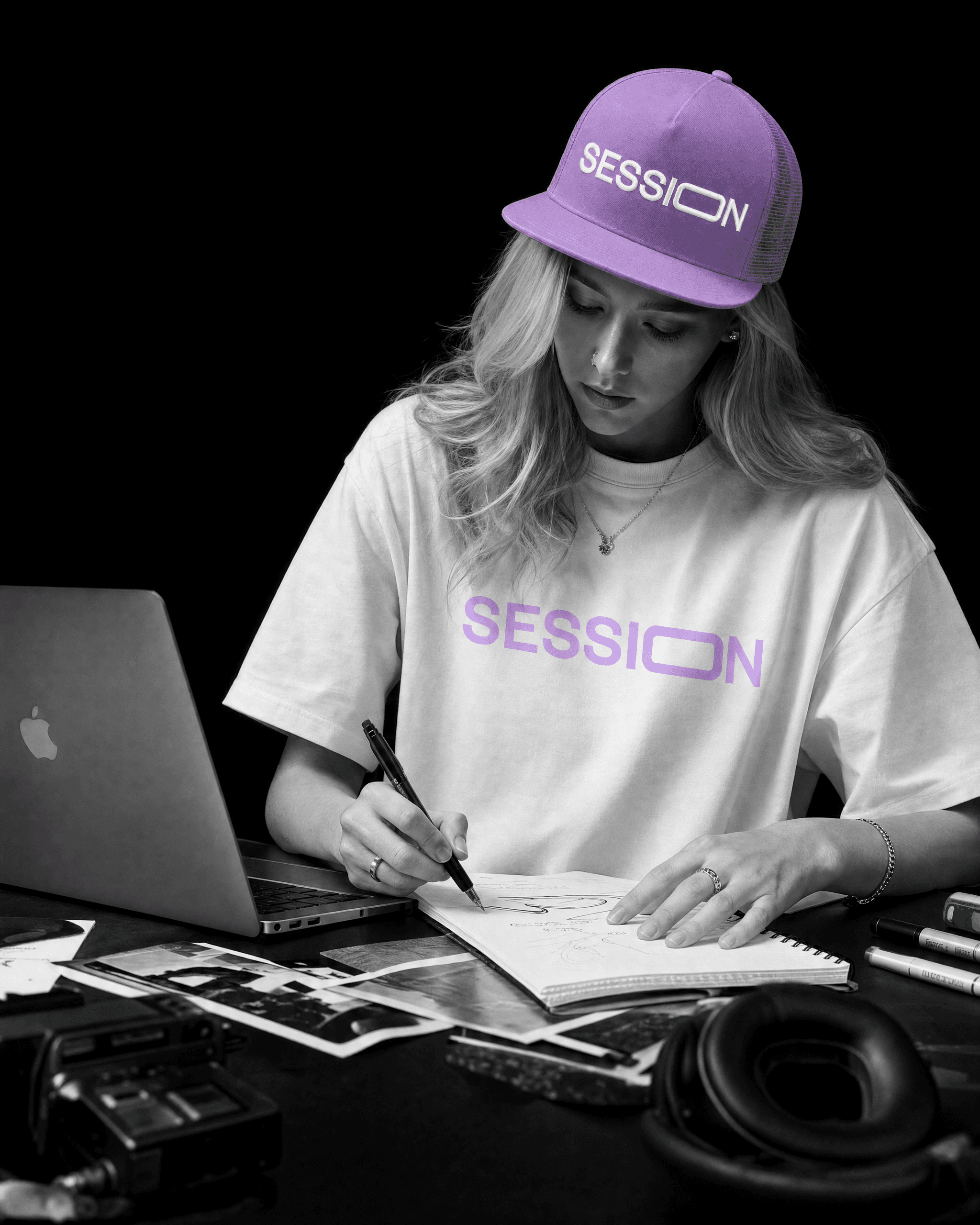 Woman in a light-purple Session Zone cap and white logo T-shirt writing beside a laptop and headphones.