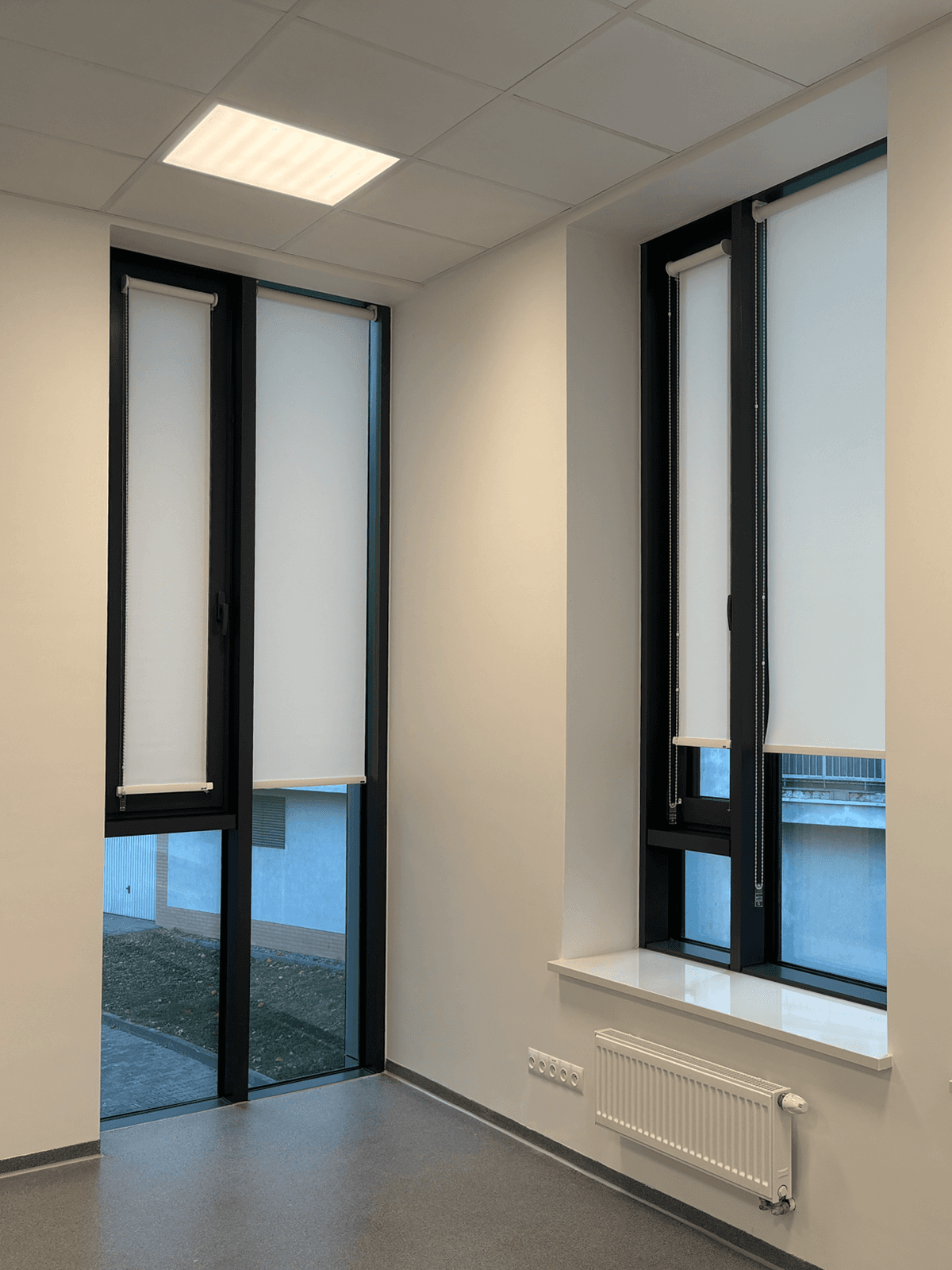 Białe rolety materiałowe Heyshade zamontowane na wysokich, czarnych oknach w nowoczesnym biurze w Pruszkowie.