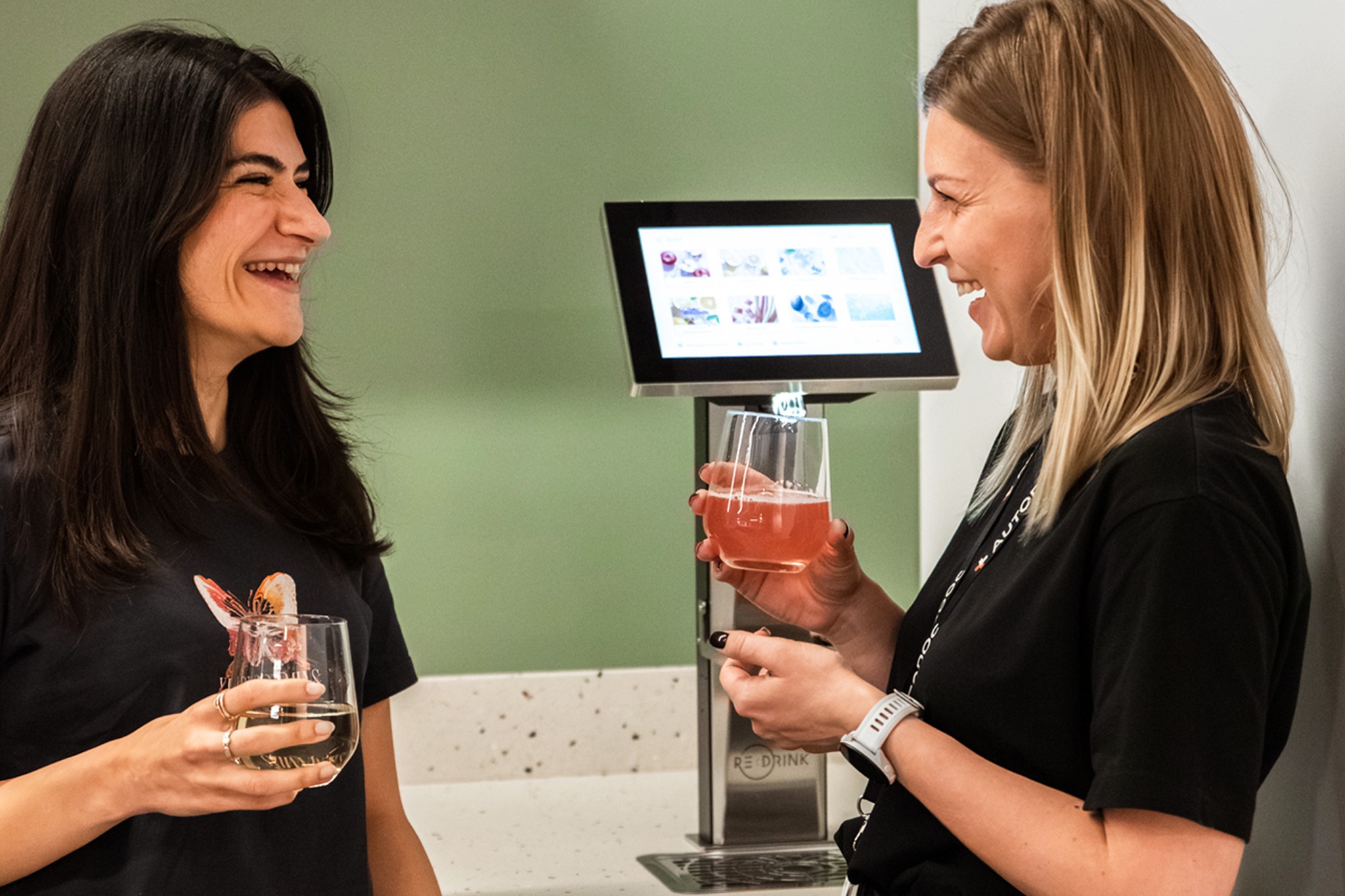 Beril and Anastasiia from AUTODOC in front of ReDrink Dispenser, holding fresh drinks and laughing