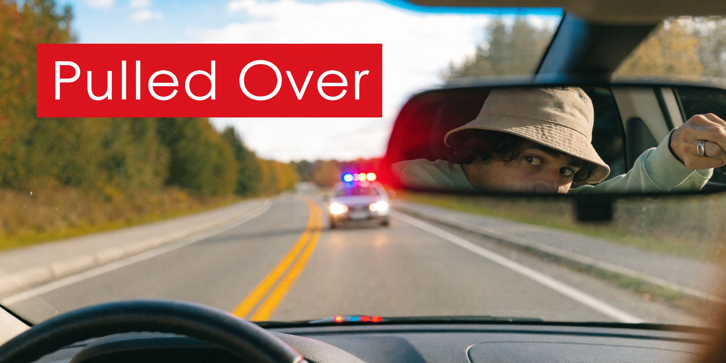 A person wearing a bucket hat looks worried in the rearview mirror as a police car approaches.