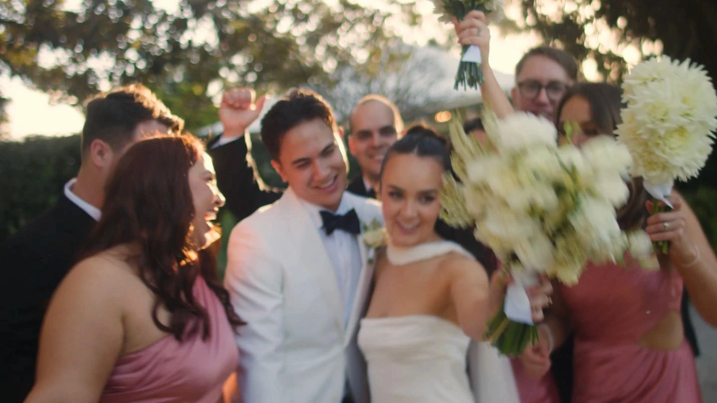 A joyful wedding party is gathered outdoors, with the bride and groom in elegant attire surrounded by friends celebrating, holding white and blush flowers amidst lush greenery and soft sunlight.