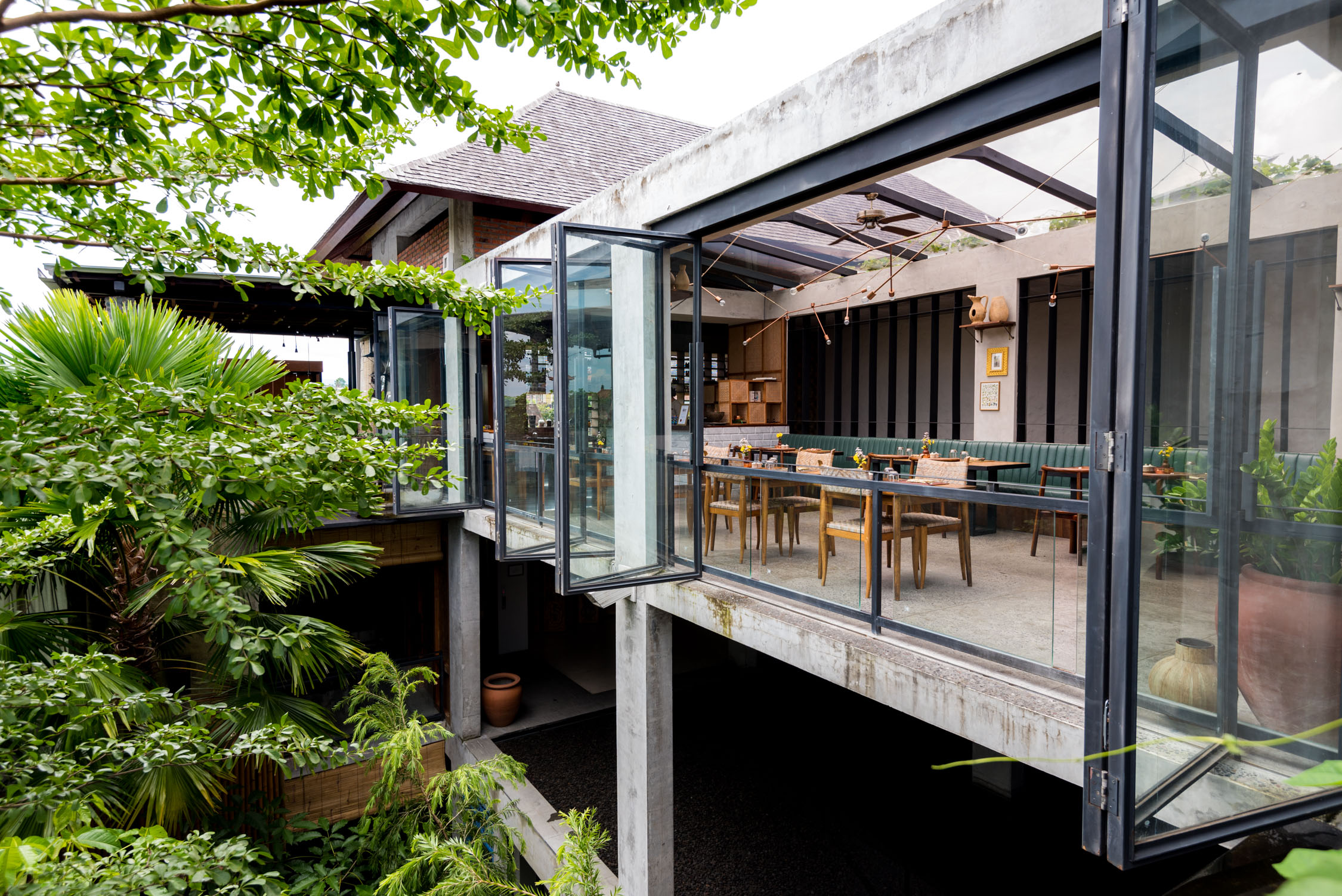 Modern concrete and glass building with open folding doors revealing a dining area with tables and chairs under trees.