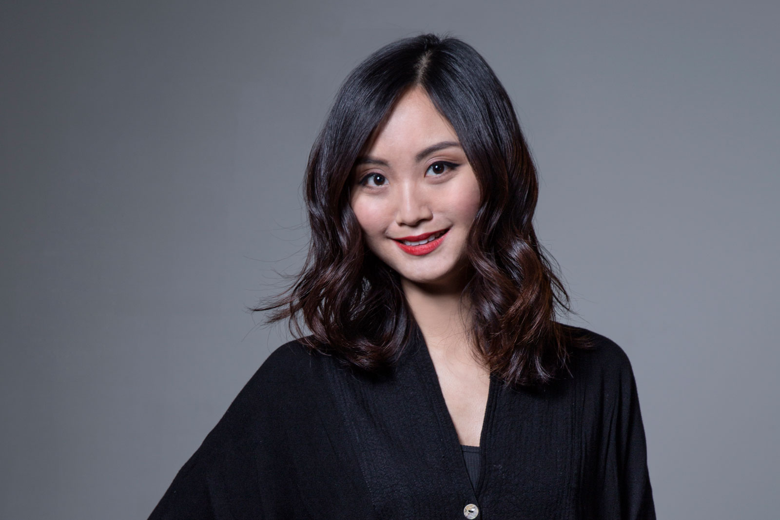 A woman with shoulder-length dark brown hair with bangs wears a black v-neck top against a solid gray background. She is looking directly at the camera with a slight smile. Her expression is neutral.