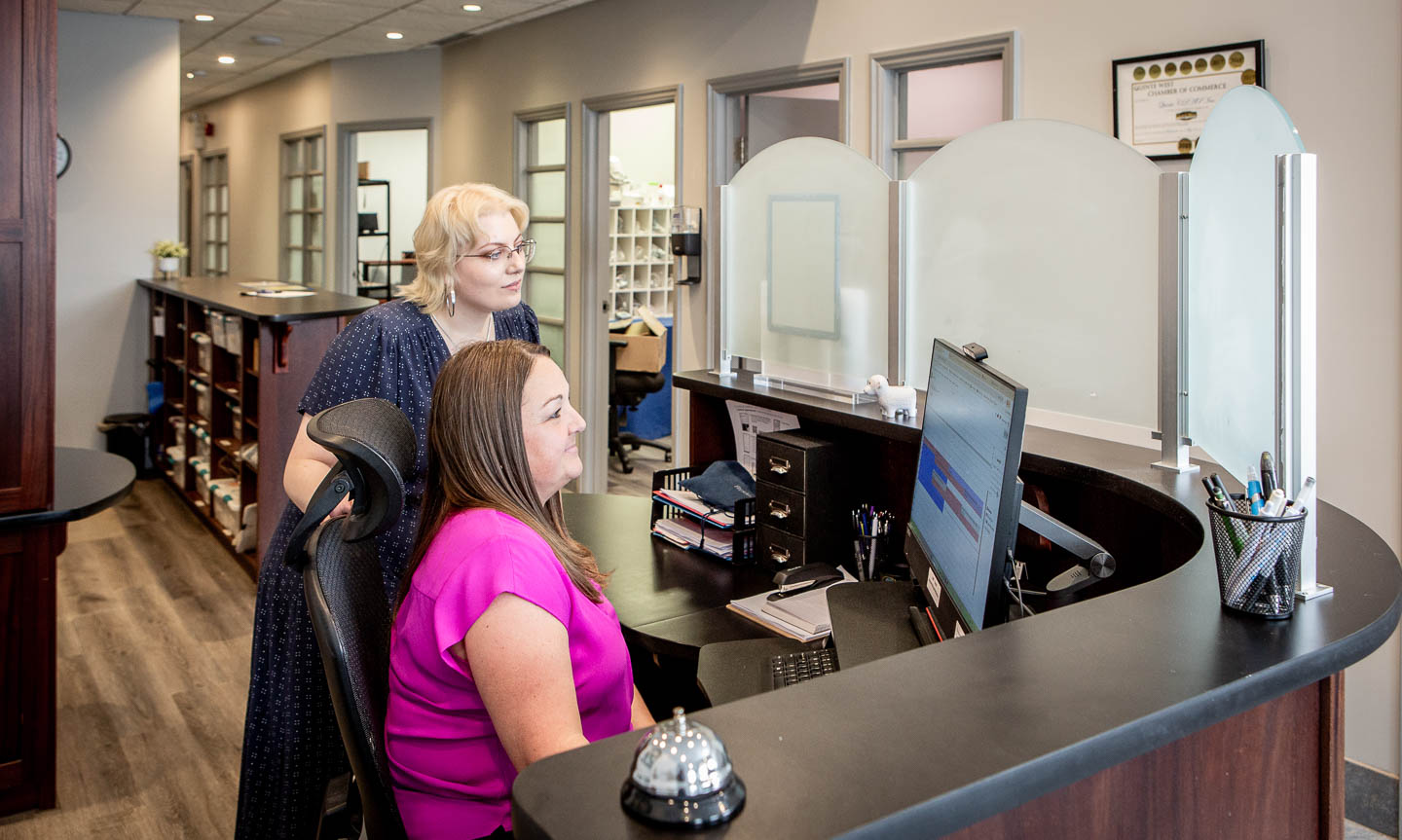 One woman sitting and one standing, both looking towards a computer screen in a CPAP clinic recpetion area.
