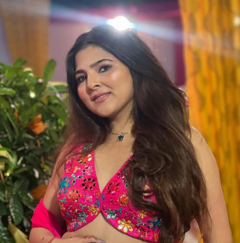 A smiling person with curly hair sits at a desk, looking at a laptop against a dark background.