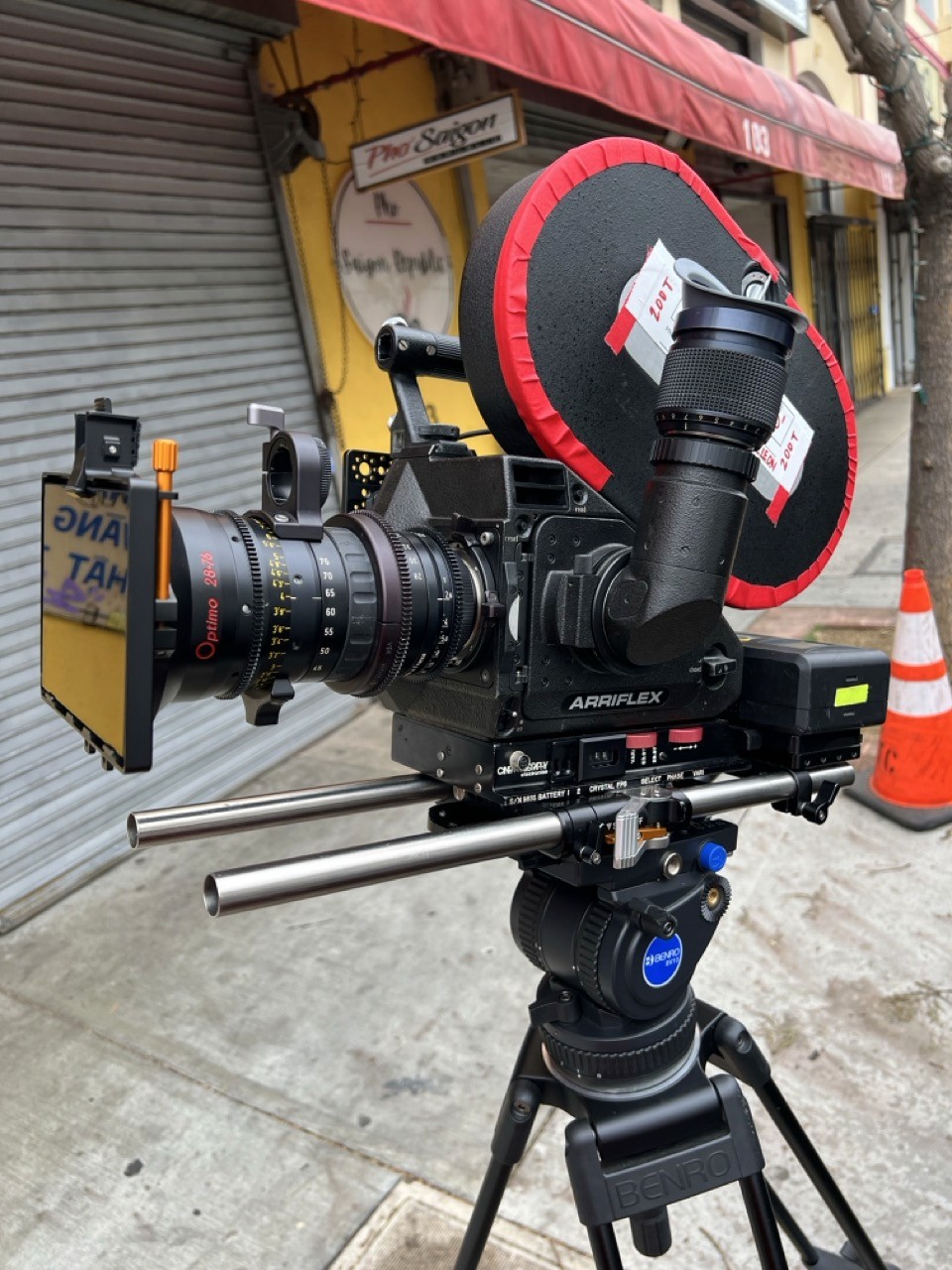 A classic Arriflex film camera with matte box and prime lens mounted on a tripod on a city sidewalk