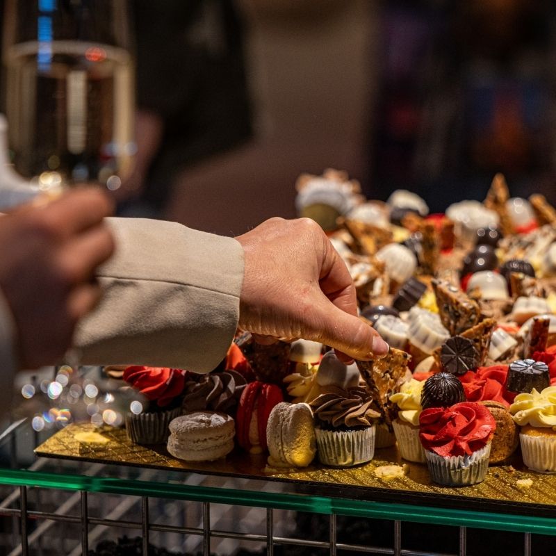 A close-up of a hand picking a dessert from a tray filled with assorted pastries, chocolates, and macarons, with a glass of sparkling drink visible in the background at a social event.