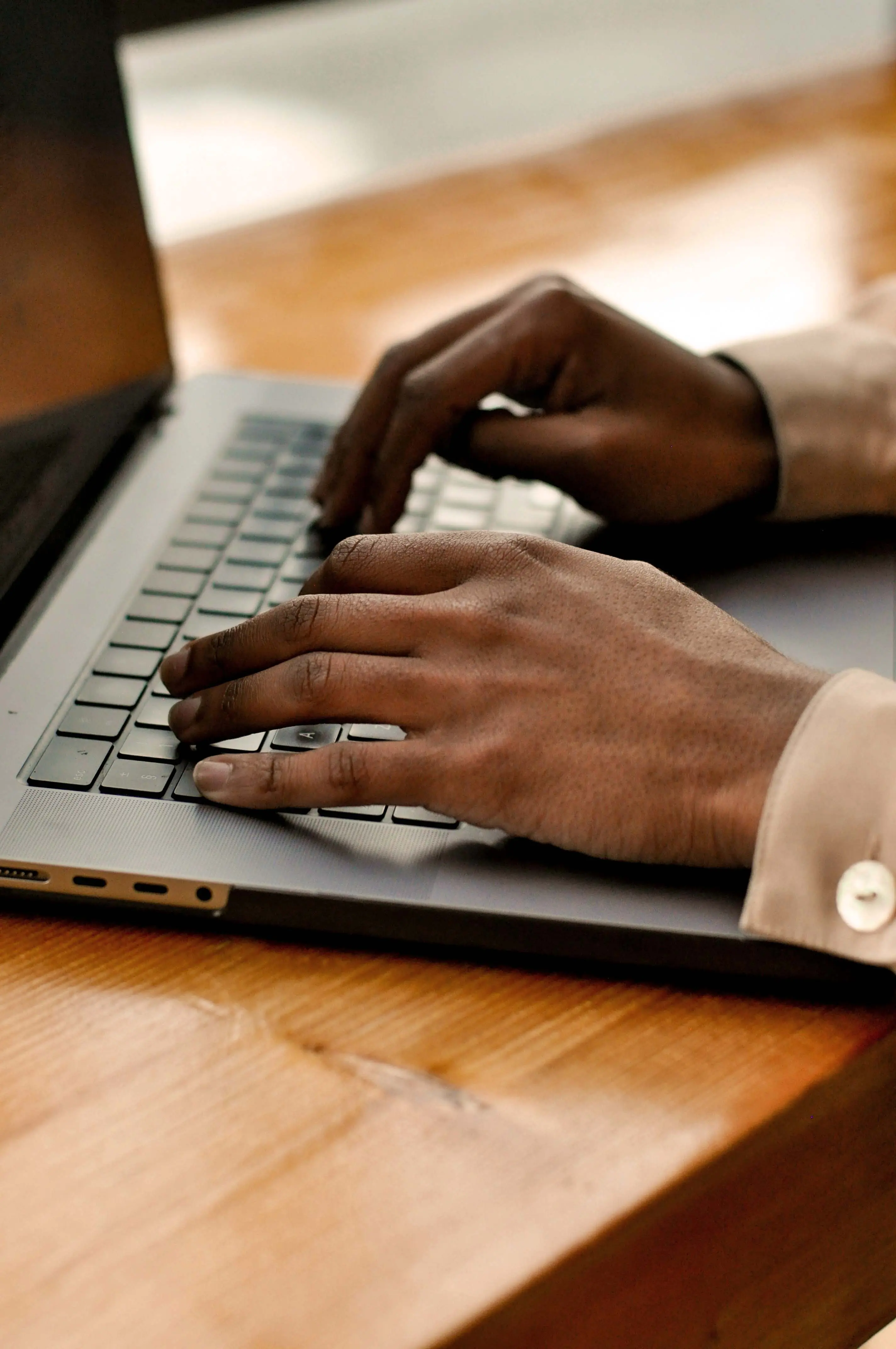 Close-up of hands typing on a laptop keyboard on a wooden desk, with one hand positioned over the keys and the other reaching toward the trackpad area.