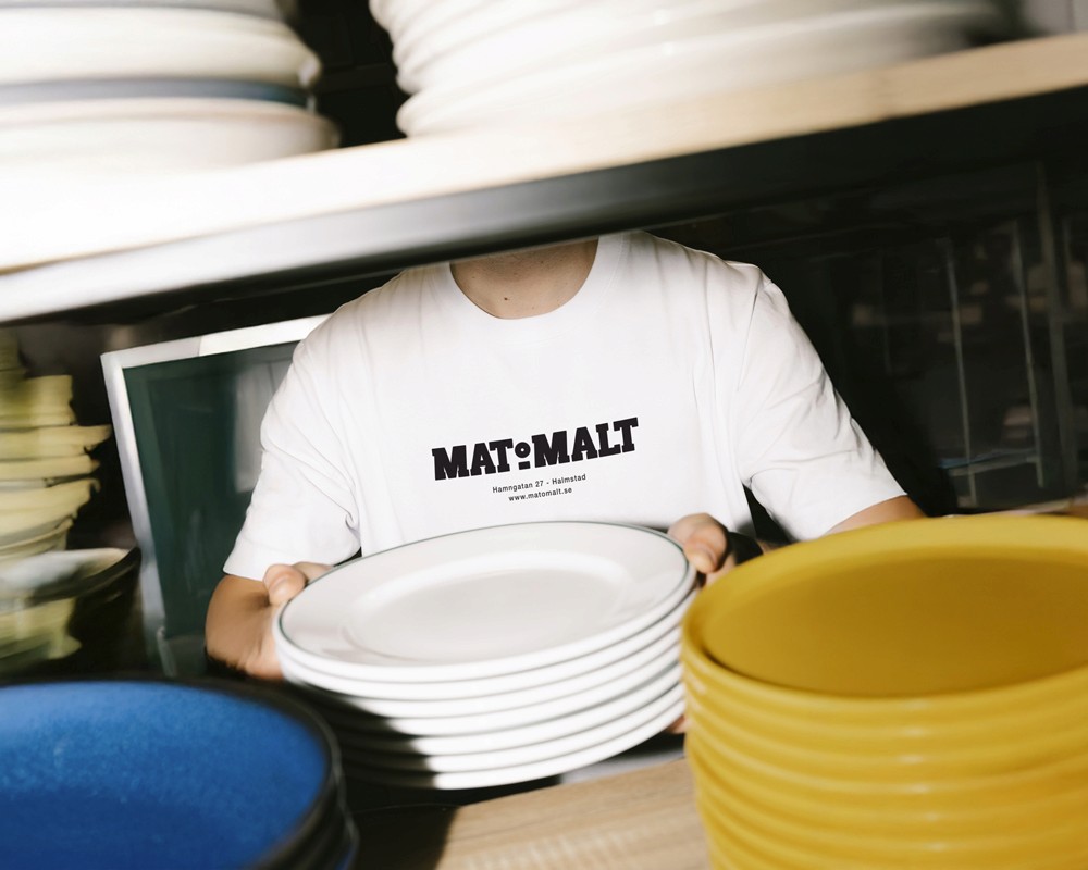 Person holding plates and wearing a white Mat & Malt branded t-shirt, demonstrating the identity applied to staff merchandise.